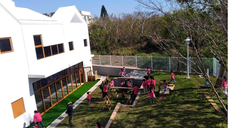 非營利幼兒園白色建築，融入綠意，小朋友遊戲其中。