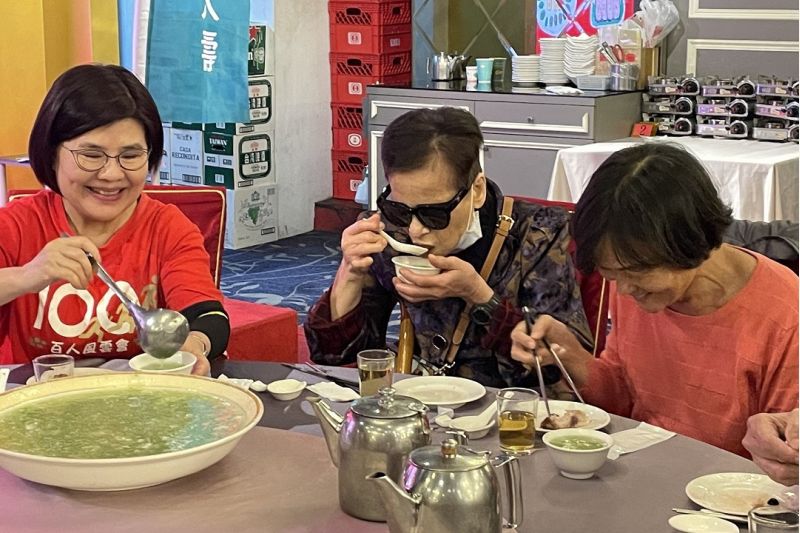 台灣人壽高三通訊處企業志工坐在旁為視障朋友舀湯。  圖片來源：尹甸基金會